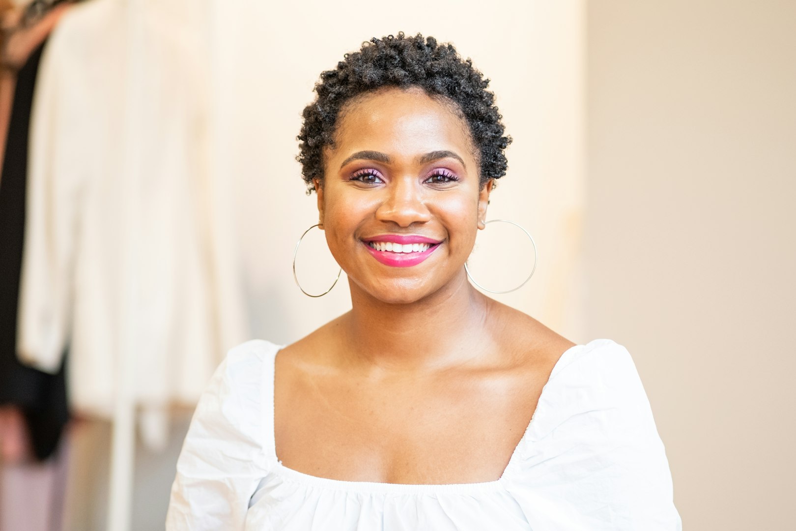 A woman with wavy, shoulder-length brown hair is smiling gently. She is wearing a white blouse and is standing in a bright, minimalist setting with turquoise decorative items in the background.